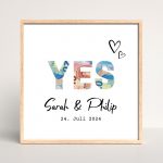Quadratisches Poster mit den Namen des Brautpaars und dem Hochzeitsdatum. Oberhalb stehen die Buchstaben "YES". Die Buchstaben wurden ausgeschnitten und hinter ihnen scheinen Geldscheine hindurch.