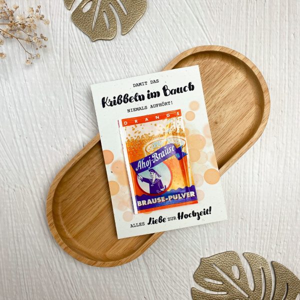 Eine ausgedruckte Karte mit einer aufgeklebten Ahoj Brause mit Geschmack Orange, im Hintergrund sind orangefarbene Blubberblasen. Auf der Karte steht der Spruch "Damit das Kribbeln im Bauch niemals aufhört. Alles Liebe zur Hochzeit".