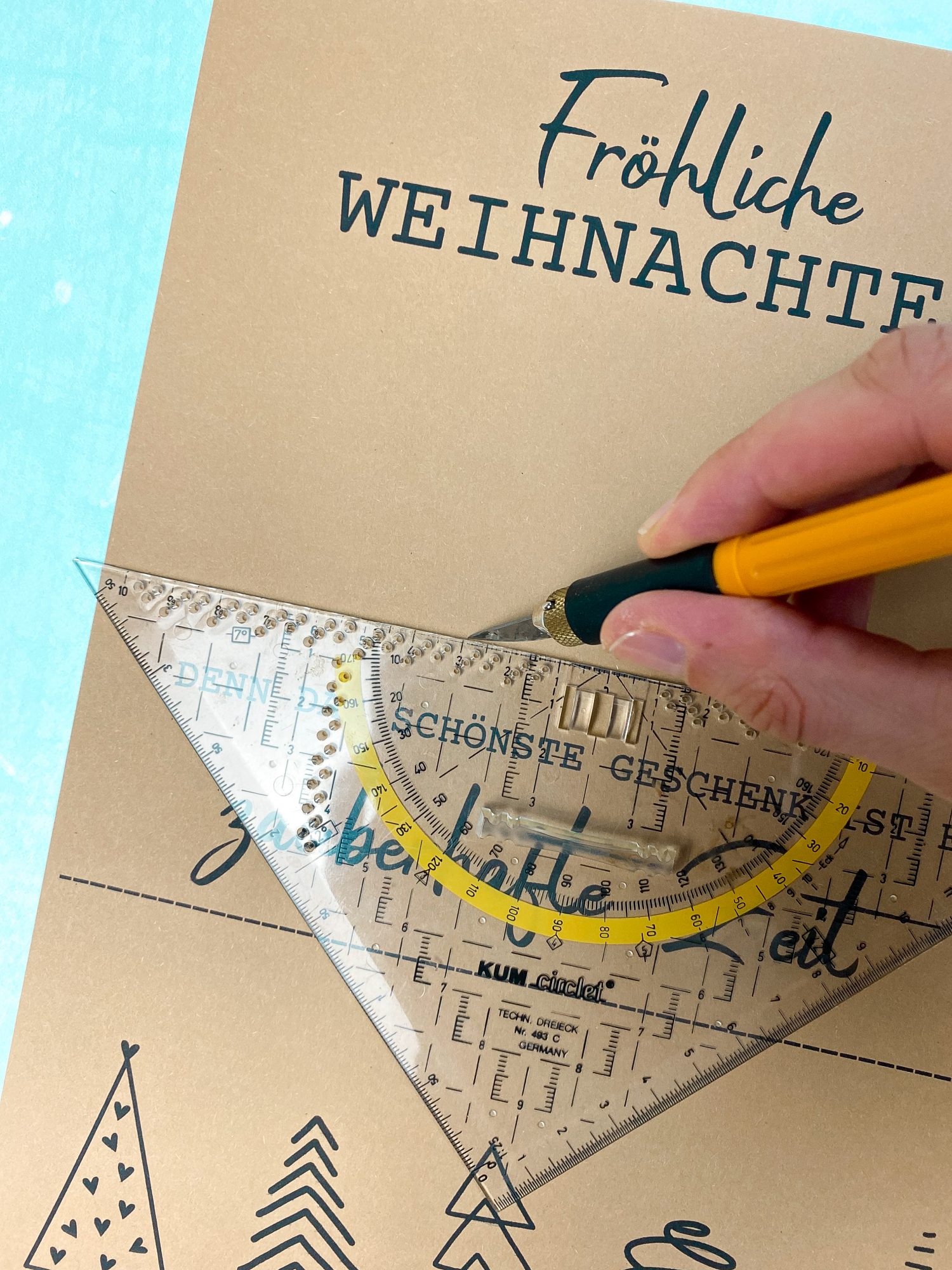 Nahaufnahme vom Basteln eines Gutscheinposters. Text „Fröhliche Weihnachten – Denn das schönste Geschenk ist eine zauberhafte Zeit“. Mit Lineal und Cutter wird ein Schlitz geschnitten, um den Gutschein einzustecken. Gutschein verpacken Weihnachten einfach selber machen.
