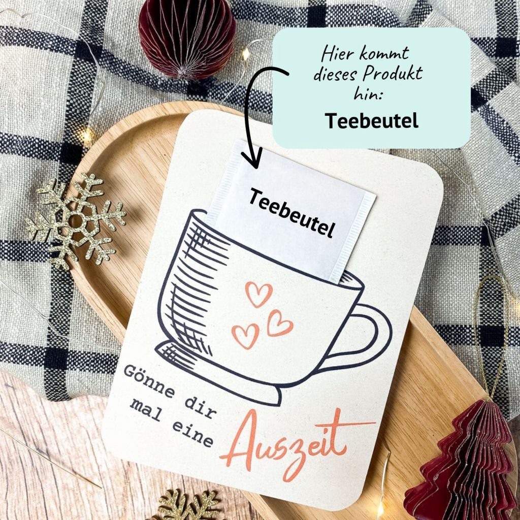 DIY Karte mit Tassenmotiv und Spruch „Gönne dir mal eine Auszeit“. In den Schlitz wird ein echter Teebeutel gesteckt. Liegt auf Holzuntergrund mit winterlicher Deko und Lichterkette – kreative Druckvorlage zum Ausdrucken für eine kleine Auszeit oder liebe Geste im Advent.