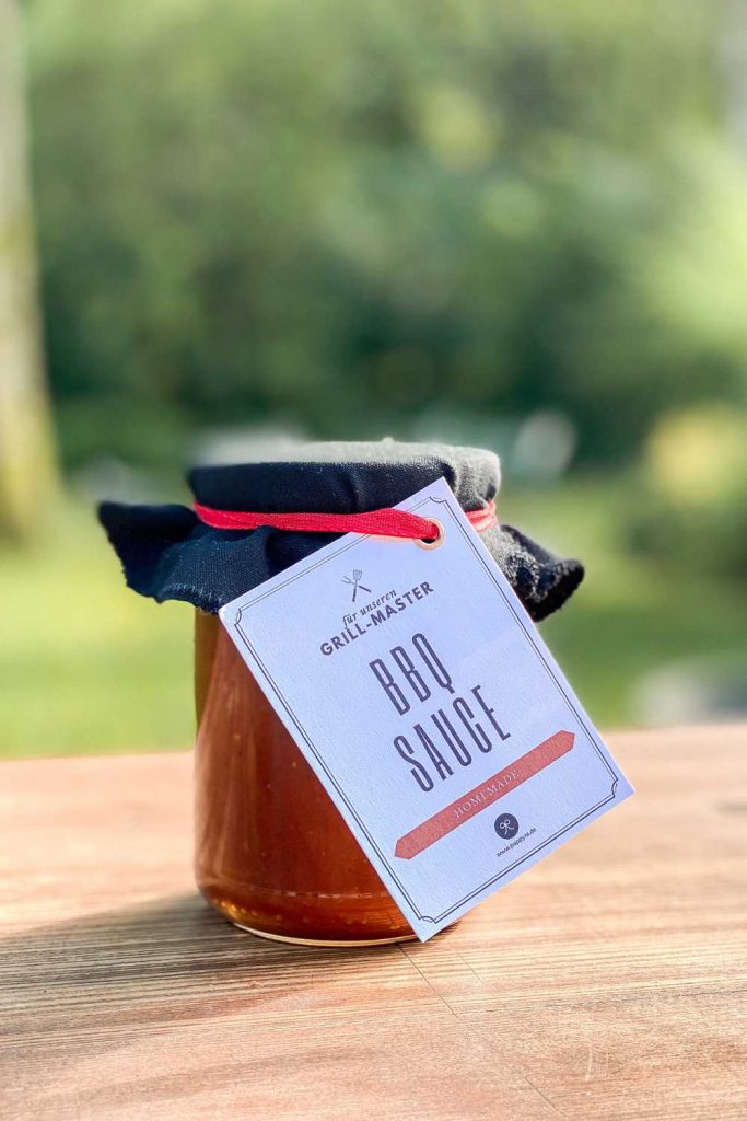 Selbstgemachte BBQ Sauce als Grillen Mitbringsel im Glas mit Etikett auf Holztisch im Garten. Perfekt als barbecue sauce selbst gemacht und ohne Zucker.