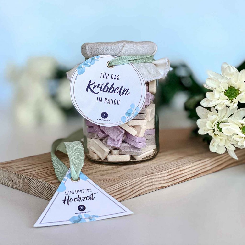 Ein liebevoll verpacktes Glas mit Stoffdeckel und Anhängern auf einem Holzbrett dekoriert mit Blumen als fertiges Beispiel für ein Geschenk zur Hochzeit DIY.