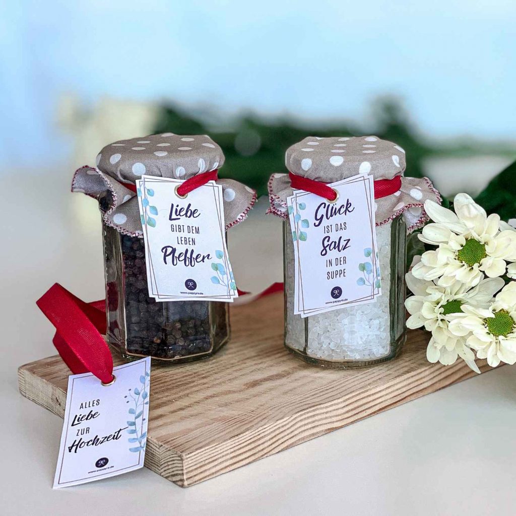 Zwei dekorierte Gläser mit Salz und Pfeffer zur Hochzeit und roten Schleifen sowie einem zusätzlichen Anhänger Alles Liebe zur Hochzeit auf einem Holzbrett.