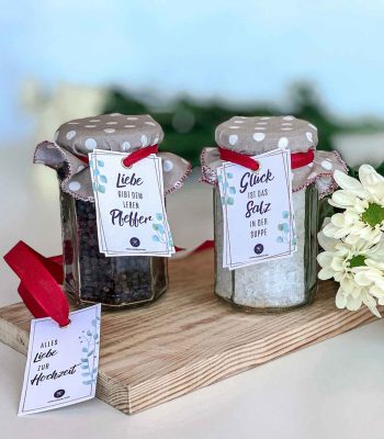 Zwei dekorierte Gläser mit Salz und Pfeffer zur Hochzeit und roten Schleifen sowie einem zusätzlichen Anhänger Alles Liebe zur Hochzeit auf einem Holzbrett.