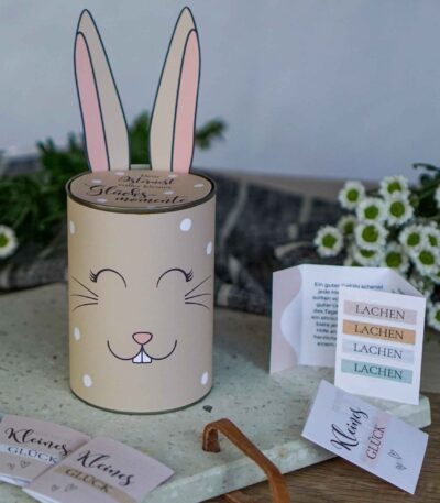 Eine beige Konservendose im Hasen-Look mit der Aufschrift Osternest voller kleiner Glücksmomente und einem geöffneten Kärtchen mit dem Wort Lachen als kreatives Ostergeschenk.