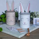 Zwei als Osterhasen dekorierte Dosen in Beige und Weiß stehen nebeneinander als Beispiel für ein kreatives Ostergeschenk mit Papierohren und kleinen Botschaften.