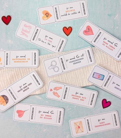 Ausgeschnittene sexy Valentinstag Gutschein Vorlagen, die dekorativ auf einem hellen Hintergrund mit kleinen Herzen verteilt sind. Die Karten zeigen verschiedene Date-Ideen wie Sex mit verbundenen Händen und Massage mit Happy End.