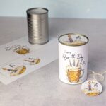 Schritt-für-Schritt Darstellung einer cleveren Bier Geburtstag Upcycling Idee. Im Hintergrund steht eine leere Konservendose samt ausgedruckter PDF Vorlage, im Vordergrund das fertige DIY Geschenk mit bedruckter Banderole, passendem Deckel-Motiv und Geschenkanhänger.
