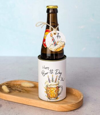 Eine braune Bierflasche wird als originelles Bier Geschenk auf einem kleinen Holztablett neben getrockneten Blumen präsentiert. Die Flasche steckt in einer Banderole mit der Aufschrift 'Happy Beer-th Day To You!' und dem Aquarell-Motiv eines Bierkrugs mit Geburtstagskerzen. Am Flaschenhals ist ein passendes, lustiges Etikett an einer Kordel befestigt. Eine perfekte Idee für ein persönliches DIY Geschenk: Einfach die digitale Druckvorlage als PDF Vorlage herunterladen, ausdrucken und im Handumdrehen ein kreatives Last Minute Geschenk basteln.
