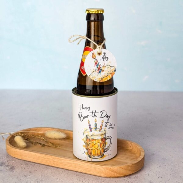 Eine braune Bierflasche wird als originelles Bier Geschenk auf einem kleinen Holztablett neben getrockneten Blumen präsentiert. Die Flasche steckt in einer Banderole mit der Aufschrift 'Happy Beer-th Day To You!' und dem Aquarell-Motiv eines Bierkrugs mit Geburtstagskerzen. Am Flaschenhals ist ein passendes, lustiges Etikett an einer Kordel befestigt. Eine perfekte Idee für ein persönliches DIY Geschenk: Einfach die digitale Druckvorlage als PDF Vorlage herunterladen, ausdrucken und im Handumdrehen ein kreatives Last Minute Geschenk basteln.
