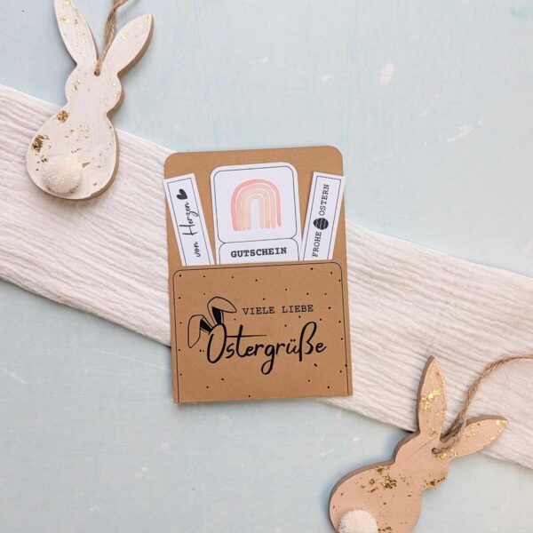Fertig gebastelter Ostern Gutschein mit dem charmanten Aufdruck "Viele liebe Ostergrüße" auf braunem Papier, dekoriert mit Holzhasen. Diese digitale Druckvorlage zum sofortigen Ausdrucken beinhaltet einen süßen Regenbogen-Gutschein und kleine Spruch-Etiketten. Eine tolle PDF Vorlage für ein schnelles, liebevolles Last Minute DIY Geschenk zum Osterfest.